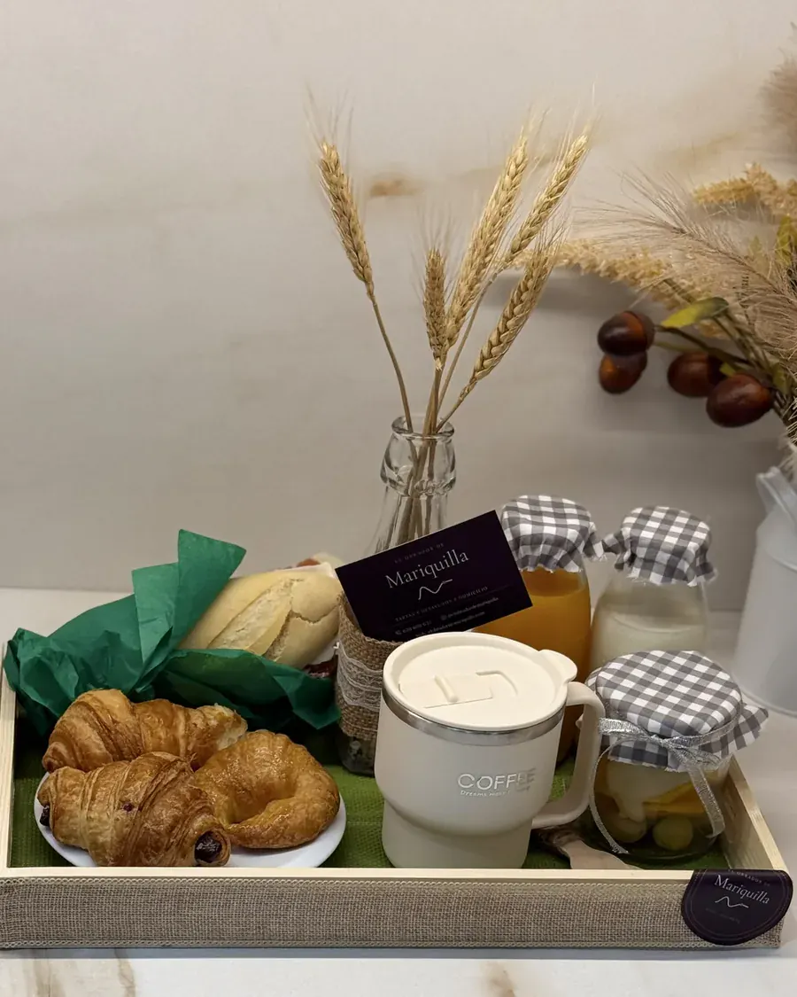 Bandeja de desayuno dulce con croissants y napolitanas, pan, taza de café, zumo, leche y fruta en tarros con tapa de cuadros, la tarjeta de Mariquilla y el sello de cierre en la esquina.