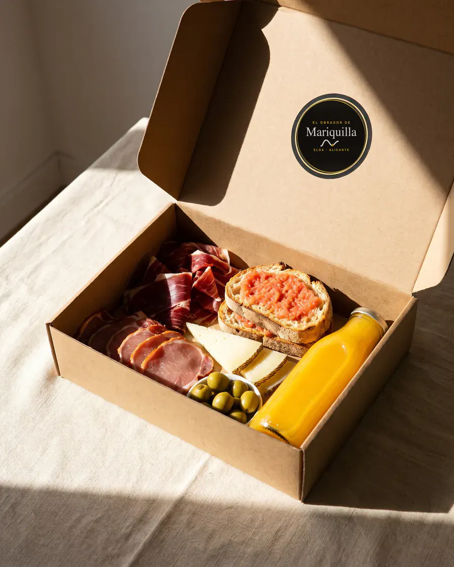 Caja de desayuno el ibérico abierta sobre mantel de lino, con el sello del obrador en la tapa.