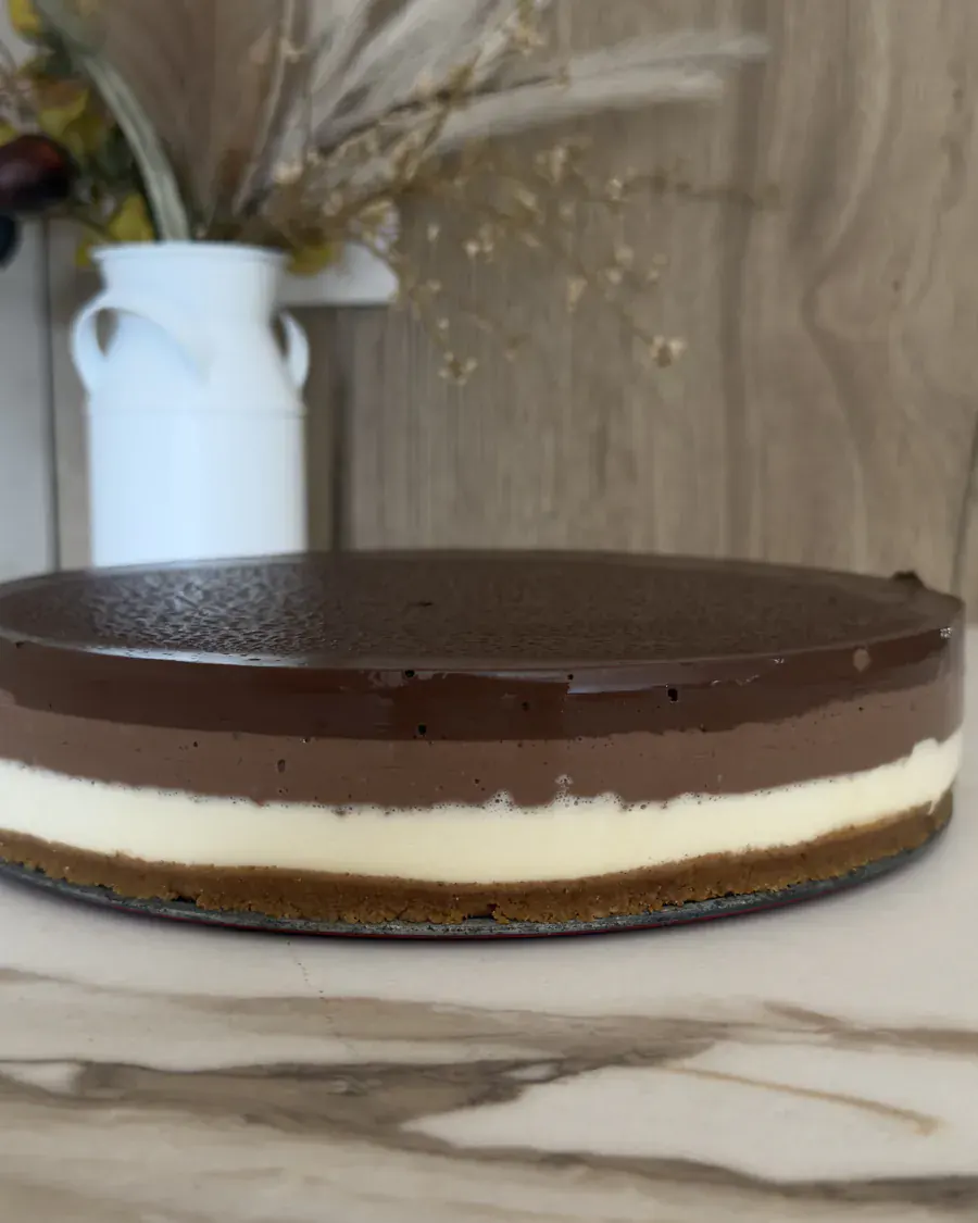 Tarta de tres chocolates entera, con sus tres capas a la vista, sobre encimera de mármol y un jarrón de flores secas detrás.