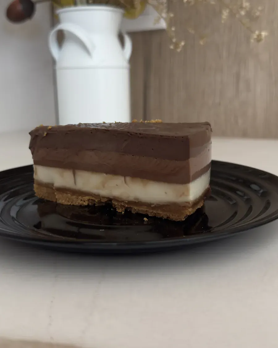 Porción de tarta de tres chocolates en un plato negro, con las tres capas y la base de galleta bien visibles.