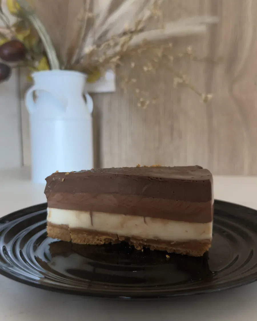Porción de tarta de tres chocolates en plato negro, con el jarrón de flores secas al fondo.
