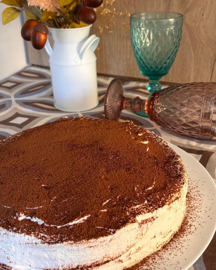 Tiramisú entero espolvoreado con cacao, con una copa de cristal verde al lado.