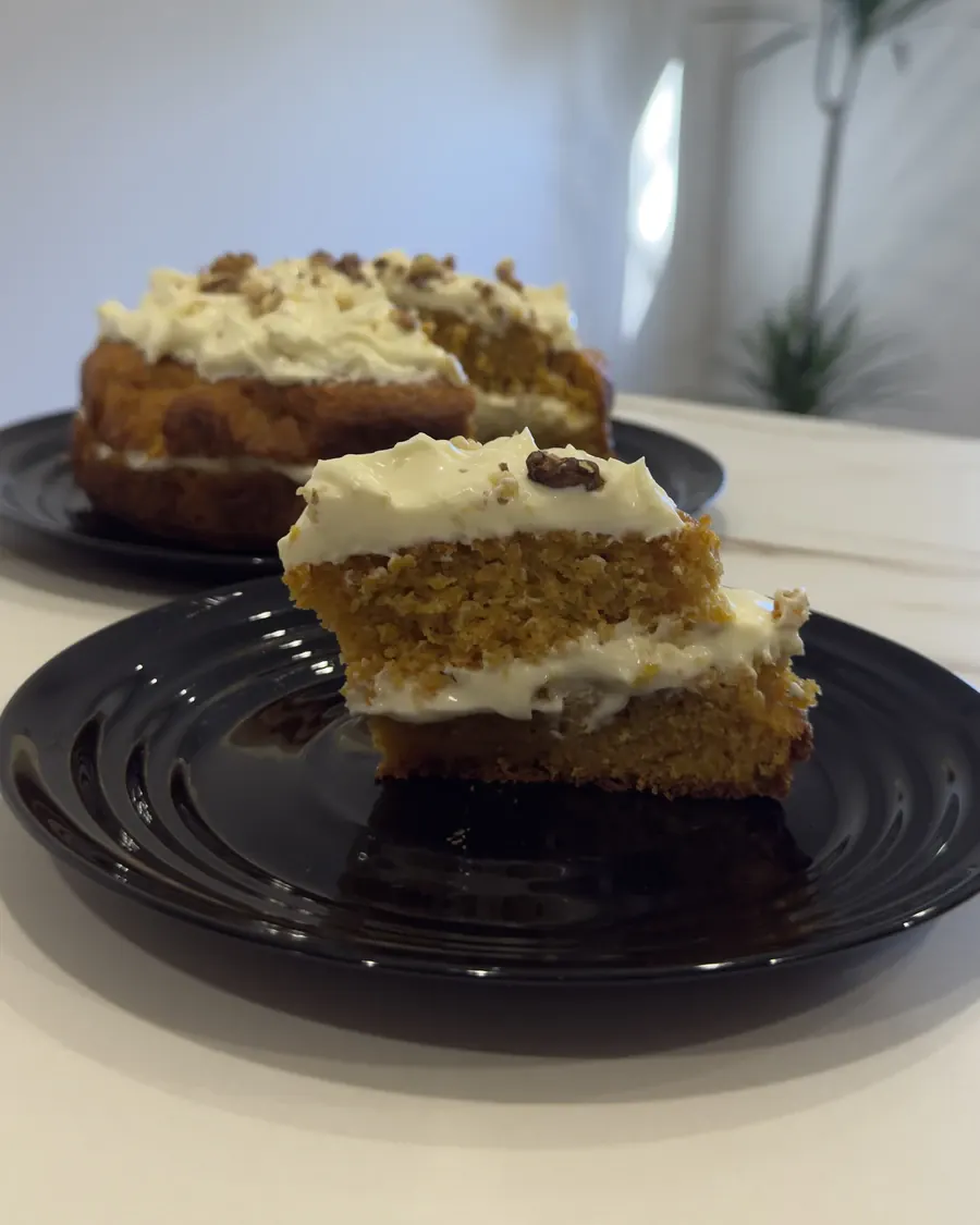 Porción de carrot cake delante de la tarta, con el bizcocho de zanahoria y la crema de queso entre capas a la vista.